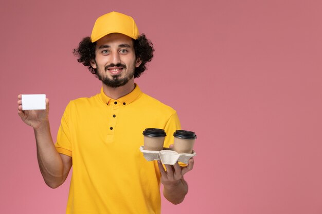 Front view male courier in yellow uniform and cape holding brown delivery coffee cups and card on pink wall