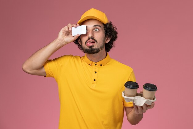 Курьер-мужчина в желтой униформе и плаще, держащий коричневые кофейные чашки и открытку на розовой стене, вид спереди