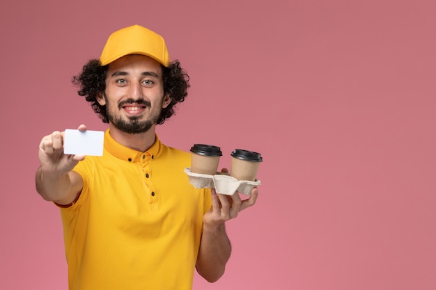 Front view male courier in yellow uniform and cape holding brown delivery coffee cups and card on pink wall