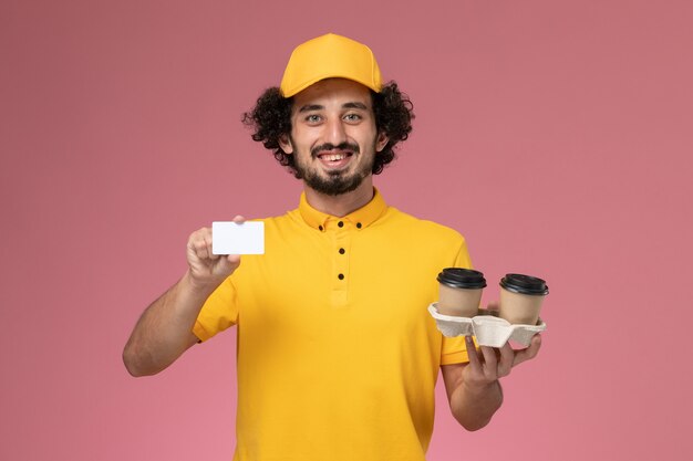 Front view male courier in yellow uniform and cape holding brown delivery coffee cups and card on the pink wall