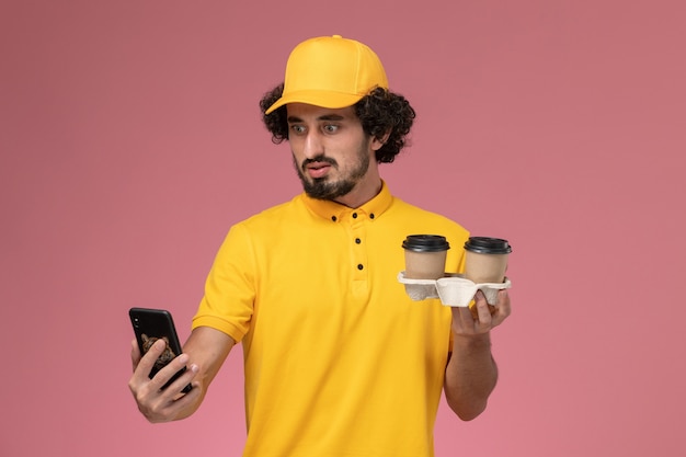 Курьер-мужчина в желтой униформе и плаще, держащий коричневые кофейные чашки, и его телефон на розовой стене, вид спереди