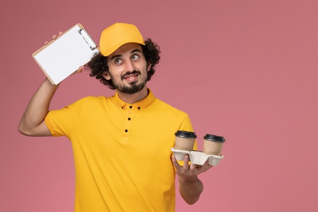 Front view male courier in yellow uniform and cape holding brown delivery coffee cups and notepad on pink wall