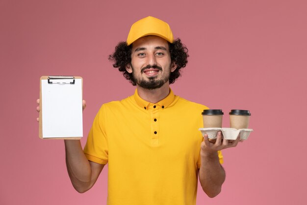 Front view male courier in yellow uniform and cape holding brown delivery coffee cups and notepad on the pink wall