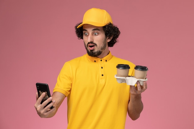 Corriere maschio di vista frontale in uniforme gialla e mantello che tiene le tazze di caffè e il telefono marroni di consegna sulla parete rosa
