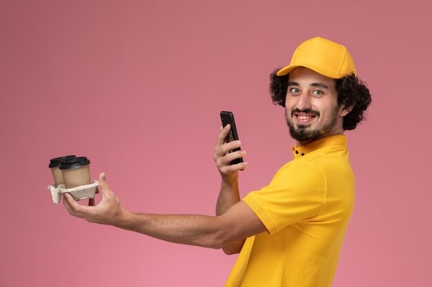 Front view male courier in yellow uniform and cape holding brown delivery coffee cups and taking photo on pink wall