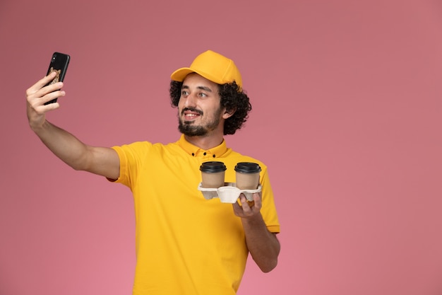 Front view male courier in yellow uniform and cape holding brown delivery coffee cups taking photo on pink wall