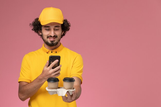 노란색 유니폼과 케이프 갈색 배달 커피 컵을 들고 분홍색 벽에 그들의 사진을 찍고 전면보기 남성 택배