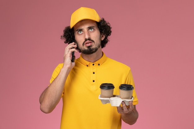 Front view male courier in yellow uniform and cape holding brown delivery coffee cups and talking on the phone on pink wall