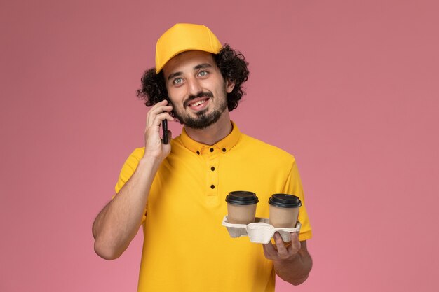 Front view male courier in yellow uniform and cape holding brown delivery coffee cups and talking on the phone on pink wall