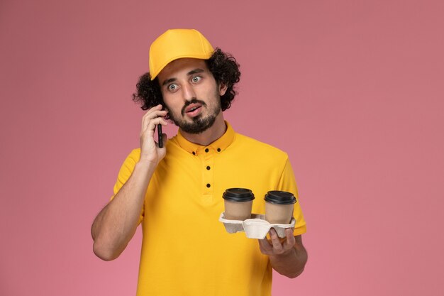 Front view male courier in yellow uniform and cape holding brown delivery coffee cups talking on the phone on pink wall