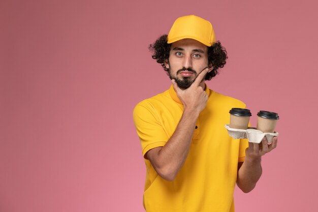 Front view male courier in yellow uniform and cape holding brown delivery coffee cups thinking on pink wall