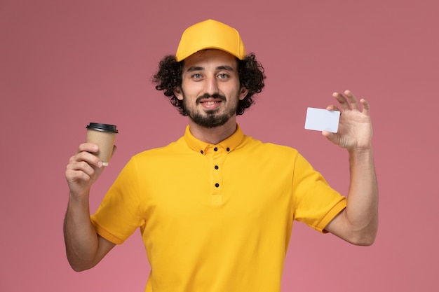 Front view male courier in yellow uniform and cape holding delivery coffee cup and card on pink wall