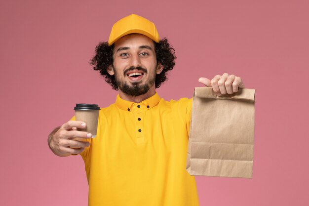 ピンクの壁に配達コーヒーカップ食品パッケージを保持している黄色の制服と岬の正面図男性宅配便