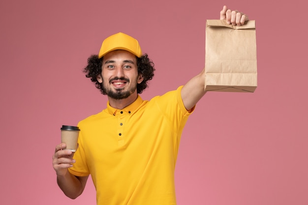 Free photo front view male courier in yellow uniform and cape holding delivery coffee cup food package on the pink wall