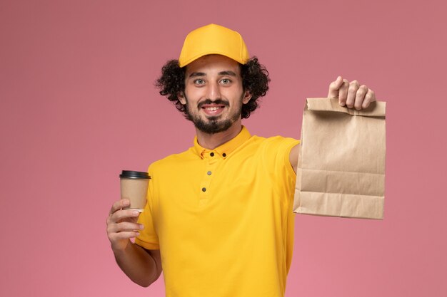 Курьер-мужчина в желтой форме и накидке с доставкой на розовой стене, вид спереди