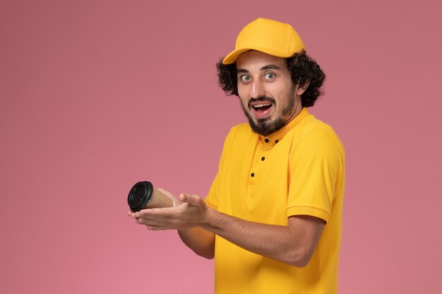 Front view male courier in yellow uniform and cape holding delivery coffee cup on light pink wall