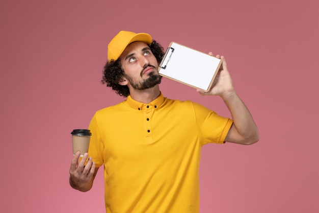 Front view male courier in yellow uniform and cape holding delivery coffee cup and notepad thinking on pink wall