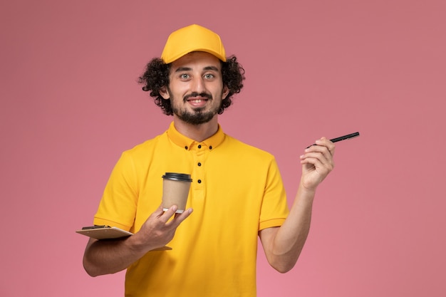 Corriere maschio di vista frontale in uniforme gialla e penna della tazza di caffè di consegna della tenuta del capo e blocco note sulla parete rosa-chiaro