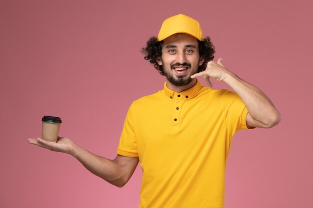 Курьер-мужчина в желтой форме и накидке, держащий чашку с доставкой кофе на розовой стене, вид спереди