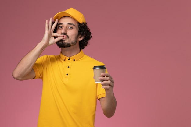 Corriere maschio di vista frontale in uniforme gialla e tazza di caffè di consegna della tenuta del capo sulla parete rosa