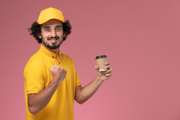 Front view male courier in yellow uniform and cape holding delivery coffee cup on the pink wall