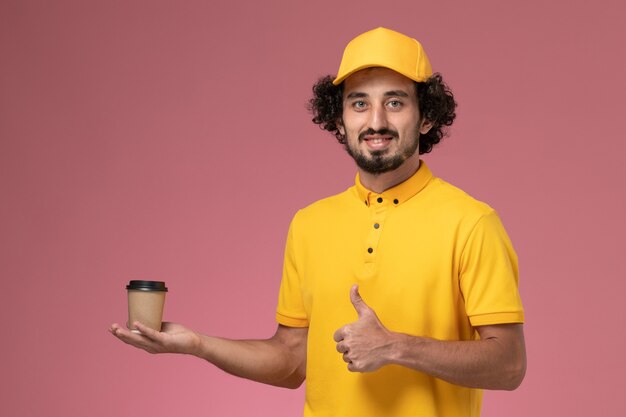 Front view male courier in yellow uniform and cape holding delivery coffee cup on the pink wall