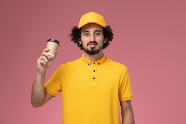 Front view male courier in yellow uniform and cape holding delivery coffee cup on the pink wall