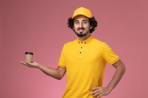 Front view male courier in yellow uniform and cape holding delivery coffee cup on the pink wall