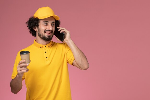 ピンクの壁に電話で話している黄色の制服とケープを保持している配達コーヒーカップの正面図男性宅配便