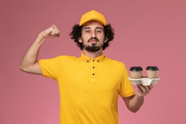 Front view male courier in yellow uniform and cape holding delivery coffee cups flexing on the pink wall