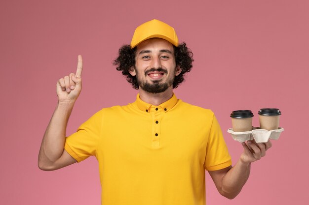 Front view male courier in yellow uniform and cape holding delivery coffee cups on the pink wall