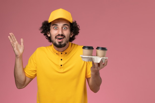 Front view male courier in yellow uniform and cape holding delivery coffee cups posing on pink wall