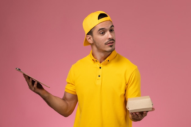 Free photo front view male courier in yellow uniform and cape holding little delivery food package and notepad on light pink background.