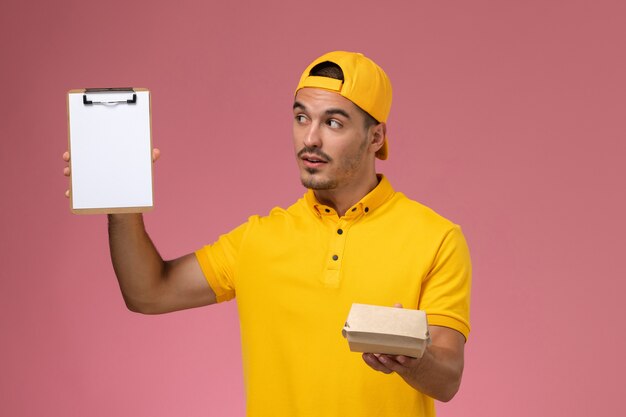 ピンクの背景に小さな配達食品パッケージのメモ帳を保持している黄色の制服と岬の正面図男性宅配便。