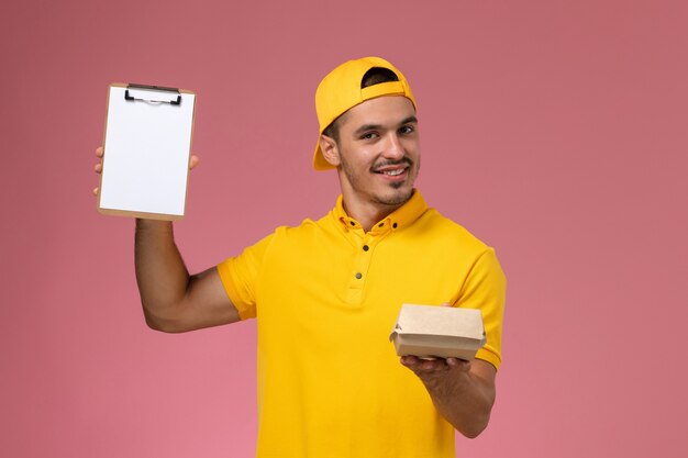 Front view male courier in yellow uniform and cape holding little delivery food package and notepad on pink background.
