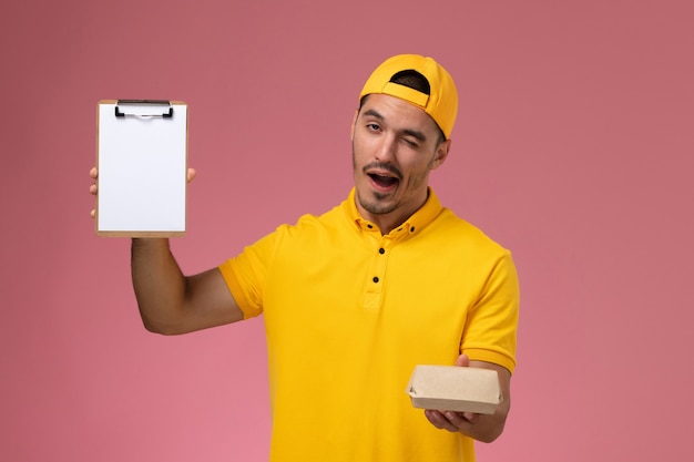 Front view male courier in yellow uniform and cape holding little delivery food package and notepad winking on pink background.