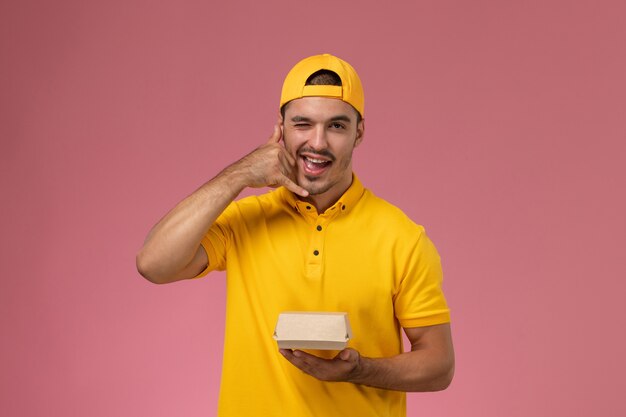 Front view male courier in yellow uniform and cape holding little delivery food package on pink background.
