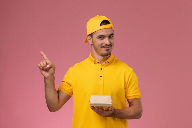 Front view male courier in yellow uniform and cape holding little delivery food package on pink background.