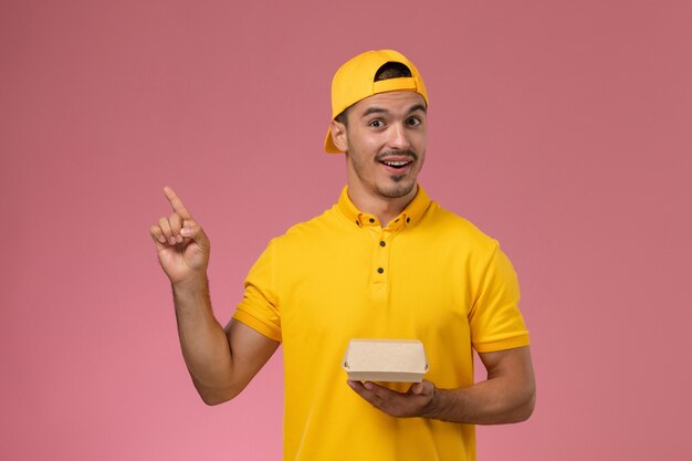 Курьер мужского пола вида спереди в желтой форме и накидке держит небольшой пакет еды доставки на розовом фоне.