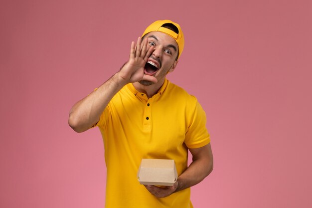 Front view male courier in yellow uniform and cape holding little delivery food package on pink background.