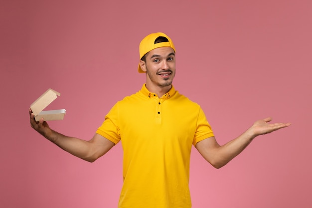 Free photo front view male courier in yellow uniform and cape holding little delivery food package on pink background.
