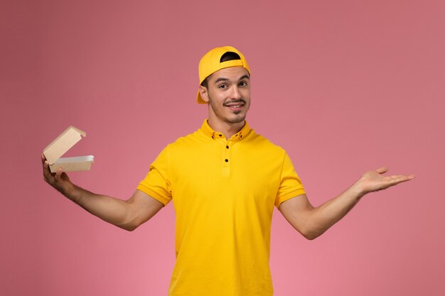 Front view male courier in yellow uniform and cape holding little delivery food package on pink background.