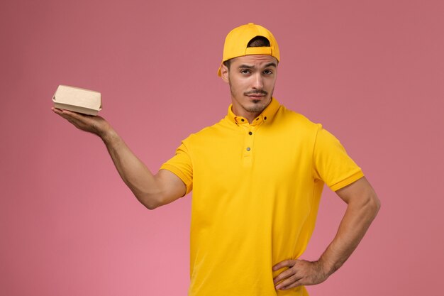 Front view male courier in yellow uniform and cape holding little delivery food package on the pink background.