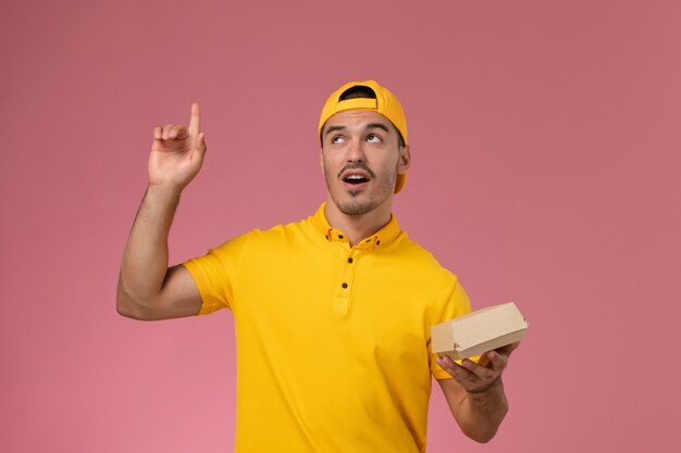 Front view male courier in yellow uniform and cape holding little delivery food package on the pink desk.