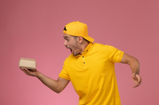 Foto gratuita corriere maschio di vista frontale in uniforme gialla e mantello che tiene piccolo pacchetto di cibo di consegna spaventato su sfondo rosa chiaro.