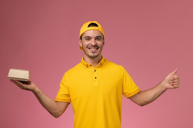 Front view male courier in yellow uniform and cape holding little delivery food package and smiling on light pink background.