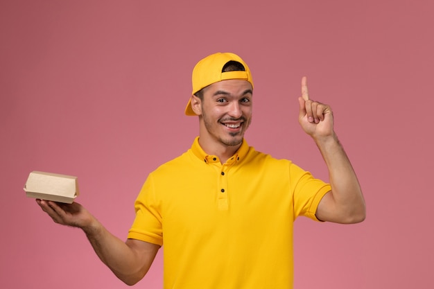 Front view male courier in yellow uniform and cape holding little delivery food package smiling on the light pink background.