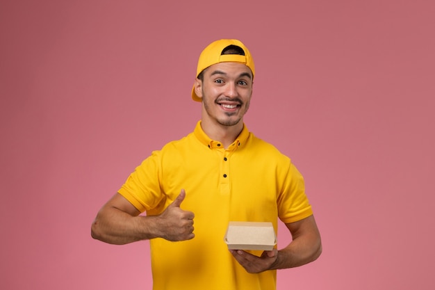 Front view male courier in yellow uniform and cape holding little delivery food package with smile on pink background.