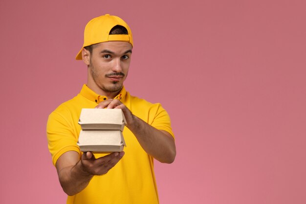 Front view male courier in yellow uniform and cape holding little delivery food packages on pink background.