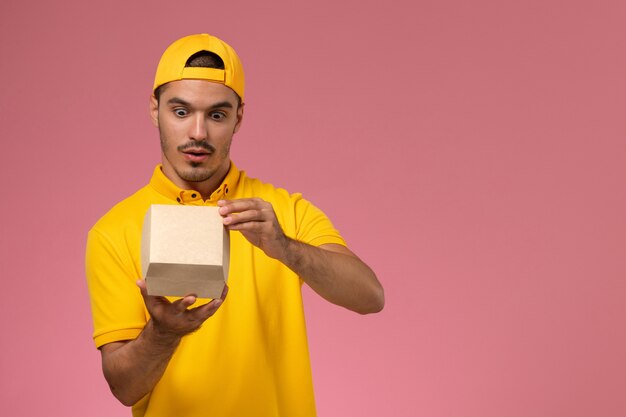 Front view male courier in yellow uniform and cape holding and opening little delivery food packages on pink background.